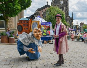 A larger than life grandma character is stood in a cobbled market town. She looks as if she has her whole world on her back. She is stoof with a costumed character, he is dressed in a calf length pink over coat and has a top hat