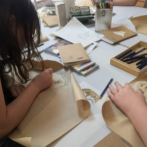 A young artists leans over her page and her hair obscures her face. On the table are drawing materials and paper and it all looks rather messy