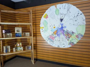 There is a large unfinished picture on the wall and next to it a set of shelves with boxes on that have been turned into mini worlds by the participants