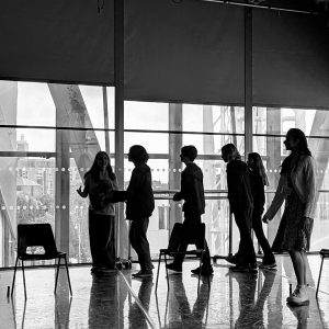 A black and white image of young people in a rehearsal room at The Lowry in Salford. There are big glass windows that frame them and the light through the window turns them into silhouettes
