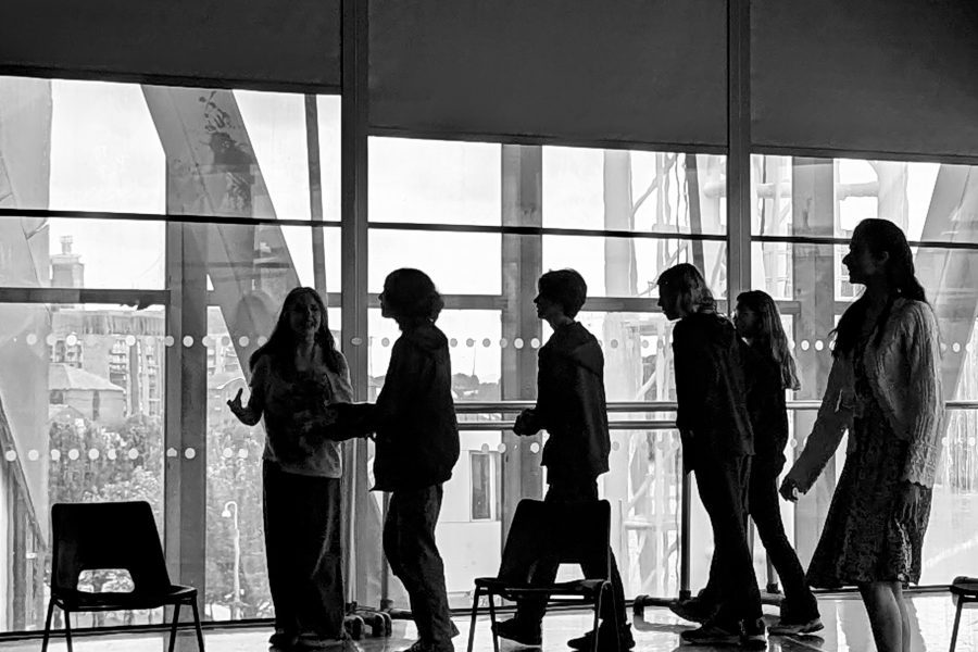 A black and white image of young people in a rehearsal room at The Lowry in Salford. There are big glass windows that frame them and the light through the window turns them into silhouettes