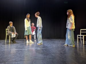 5 teenagers on stage. Three of them are standing and two are seated. Two standing are argueing with one another and the rest look on