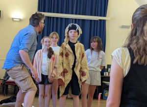 A director gets involved in a scene showing the children what he wants them to do. They are all looking at him and he has his back to the camera