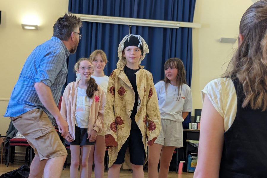 A director gets involved in a scene showing the children what he wants them to do. They are all looking at him and he has his back to the camera