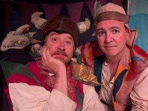 Two actors dressed up in costumes, wearing hats. They are looking at the camera and a small dragon puppet is sat on the shoulder of the man on the left