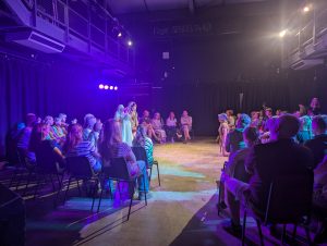A performance phot. Children are on stage and the audience surround them watching on.