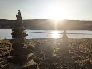 Photo of the eclipse; rocks are carefully stacked one on top of the other on the waters edge of a reservoir, the sun is low in the sky
