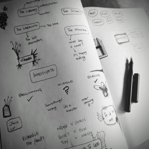 A black and white image of a notebook. Pages are open and there are drawings and words on the pages. A fountain pen lays on the page with its lid off.