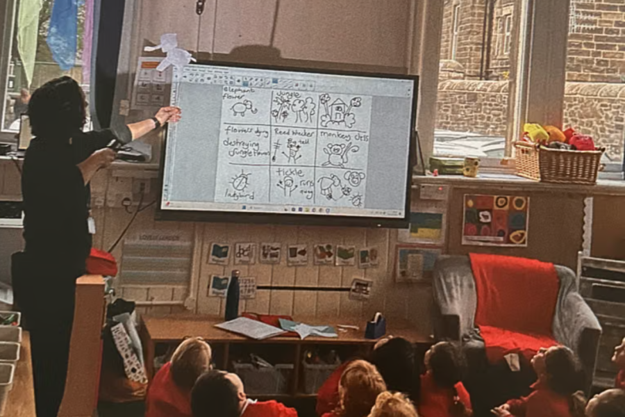 The photographer is stood behind all the children. An artist holds up a shadow puppet to a class of school children. They are all looking up at the shadow it has created