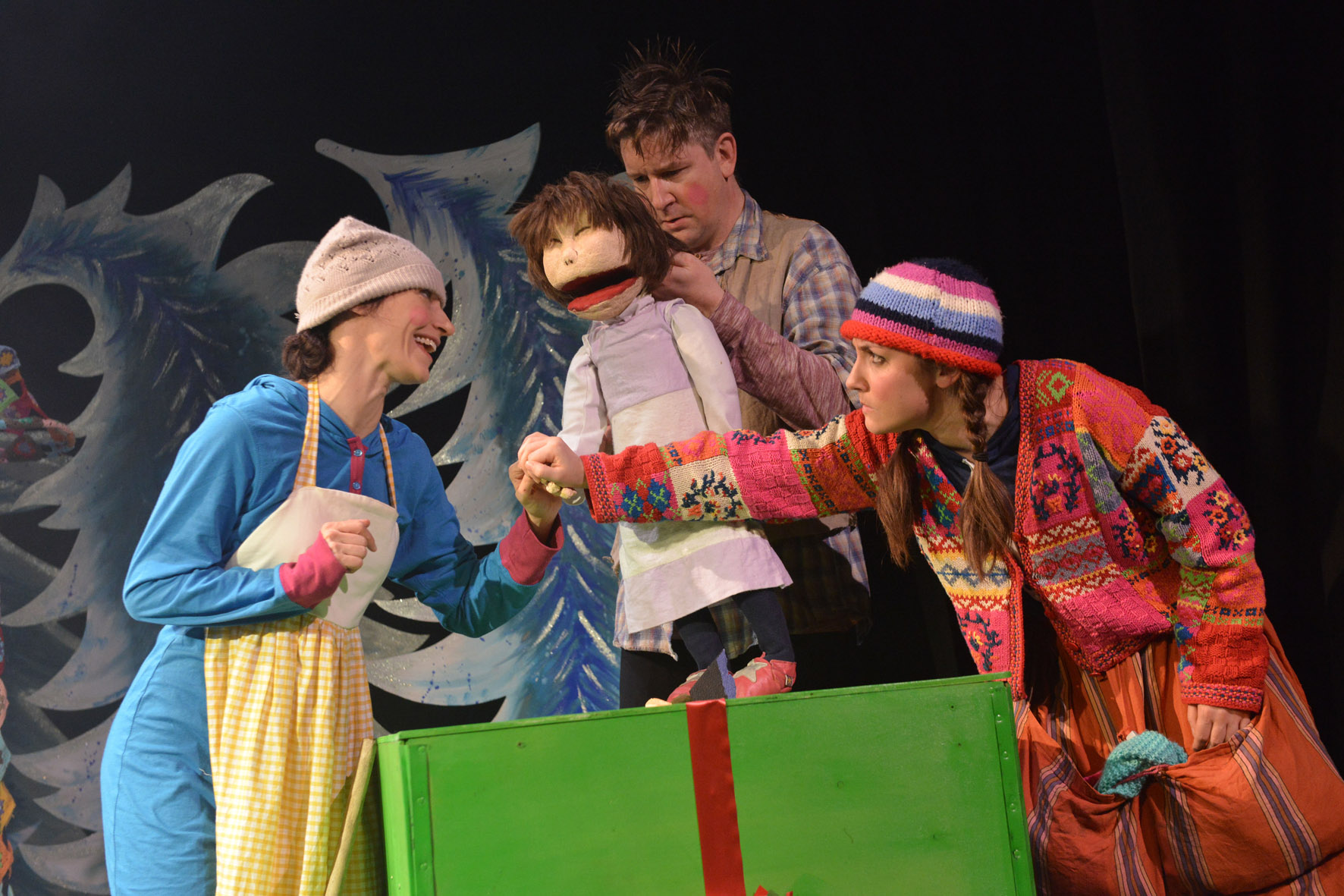 3 performers are around a green box the size of a small table. One of them is operating a puppet of a small child.