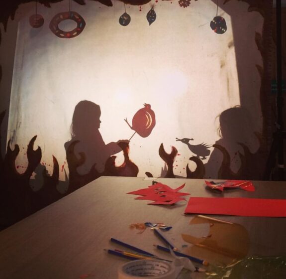 A shadow of a young girl on a white screen. Inforn of the screen is a table with all the things you need to make a shadow puppet