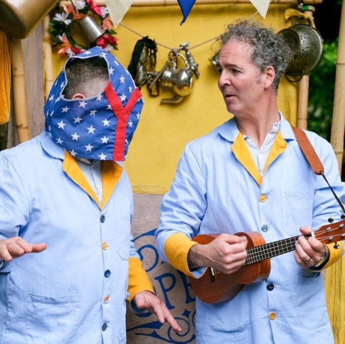 Two male performers dressed in blue stand infront of a bright yellow cart. One of them has bright blue starry pants on his head!