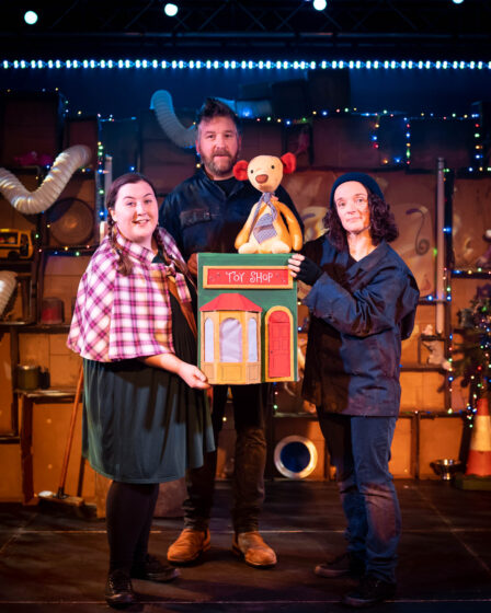 Three performers stand on stage and look out to the camera as they hold up a play toyshop and an old teddy.